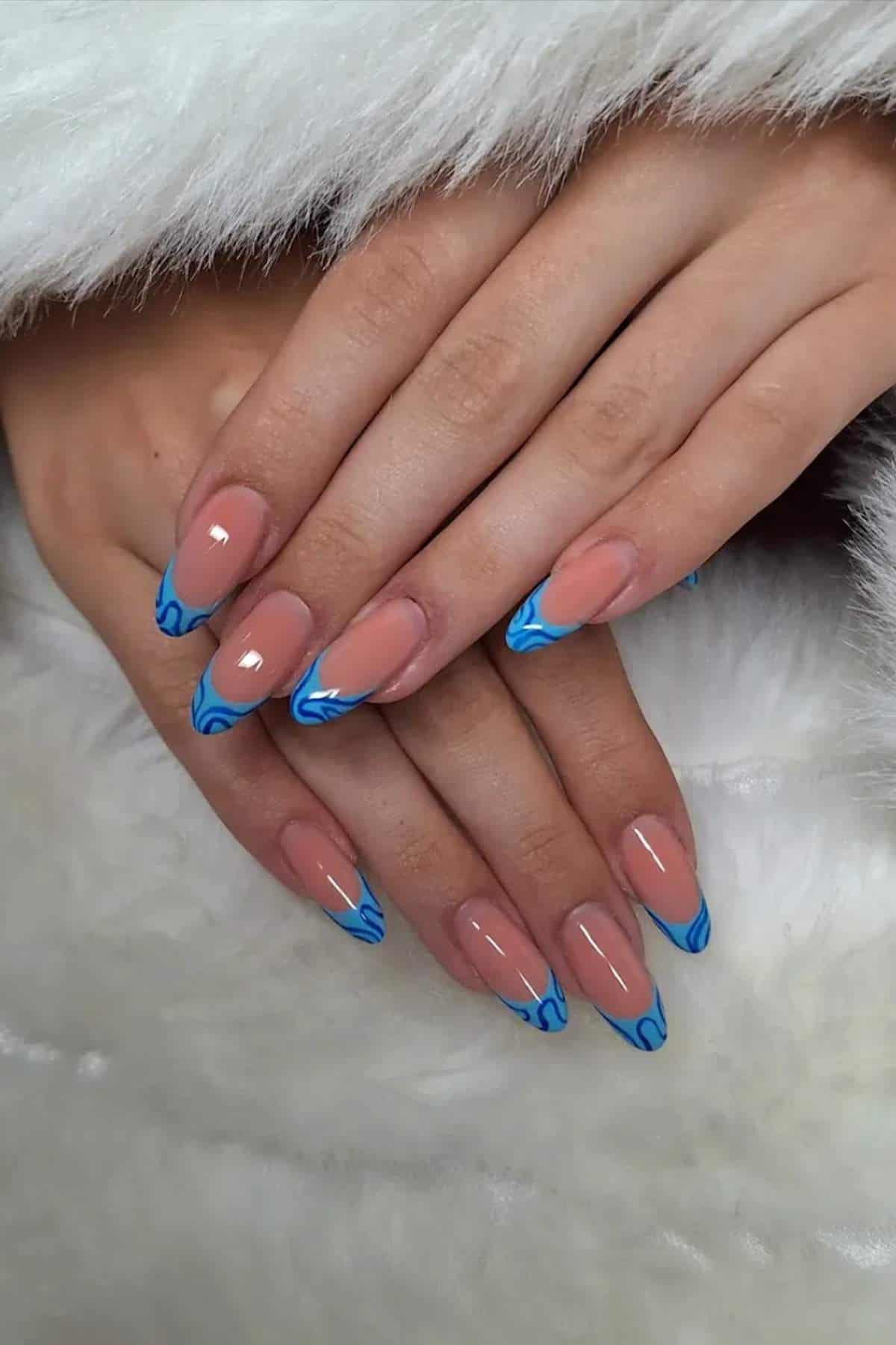 Hands with almond-shaped nails featuring a nude base and blue flame designs on the tips, resting on a white furry surface.