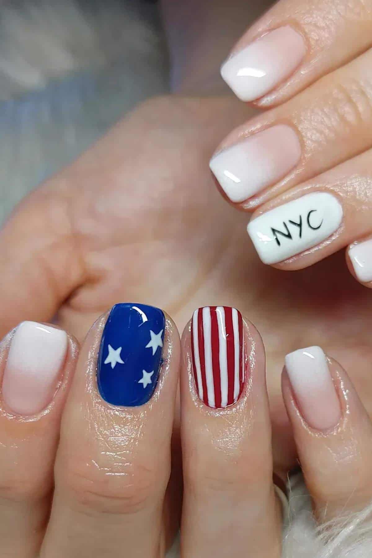 A hand with a French ombre manicure; one nail says "NYC," another has a blue background with white stars, and another has red and white vertical stripes.