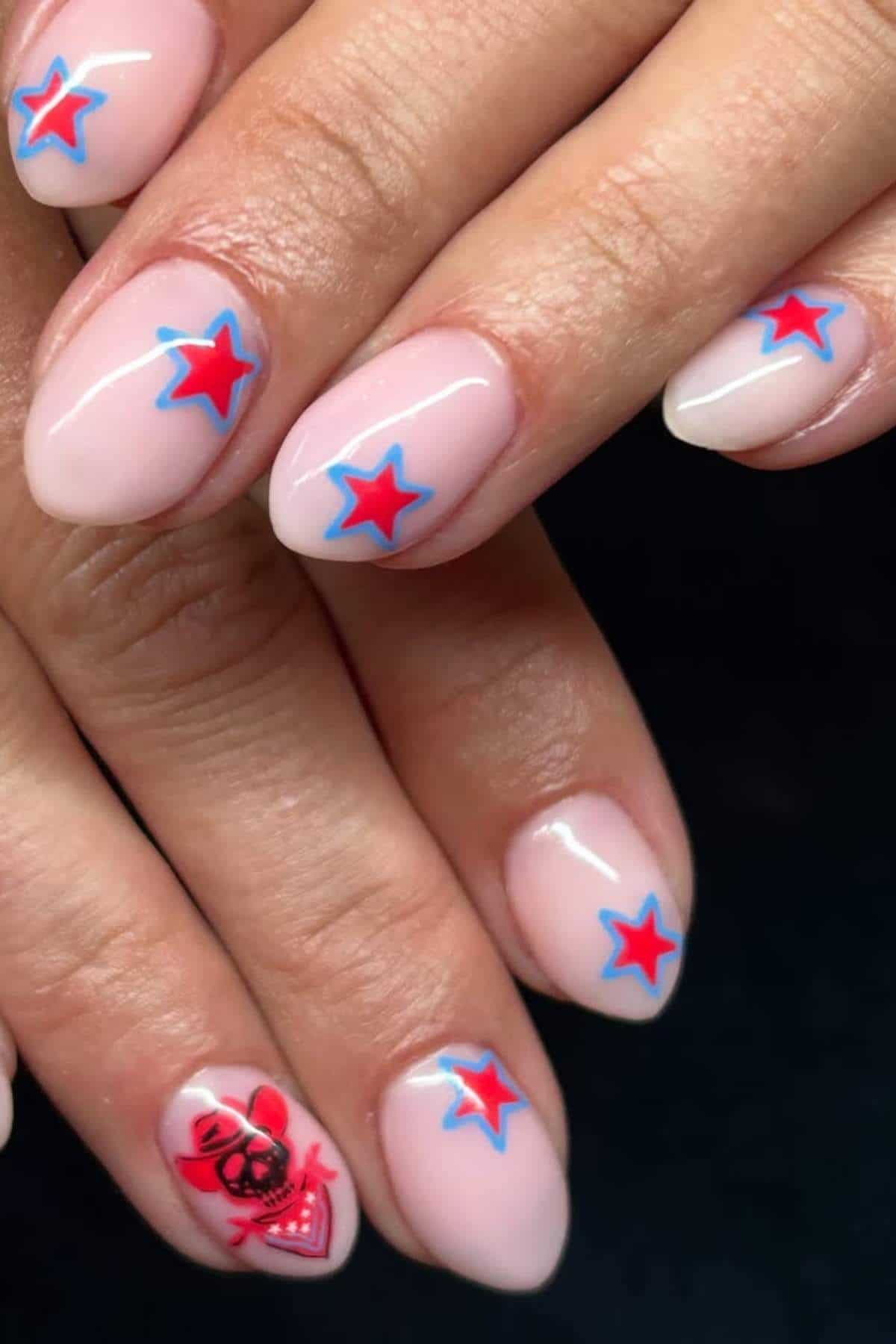 Fingernails with a light pink base, decorated with red and blue stars on most nails and a red skull with a bow on one nail.