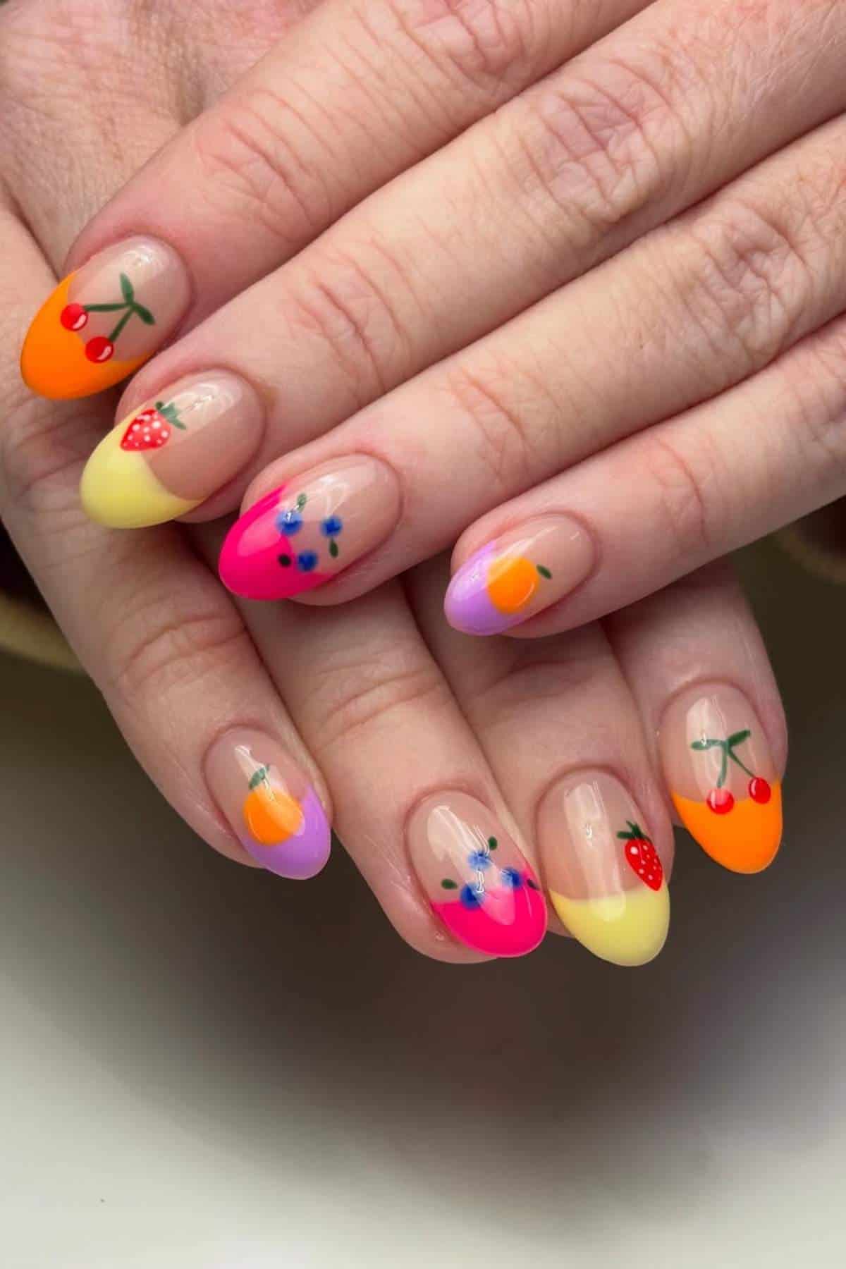 Close-up of hands with almond-shaped nails featuring colorful French tips and small fruit designs, including cherries, strawberries, and blueberries on a nude base.