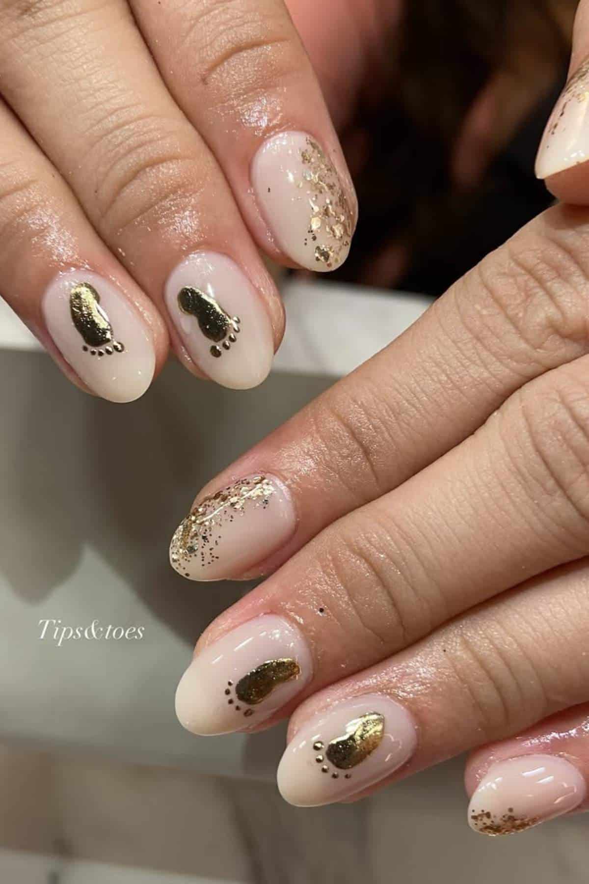 Hands with almond-shaped nails featuring nude polish, gold foil accents, and small gold footprint designs on the ring fingers.