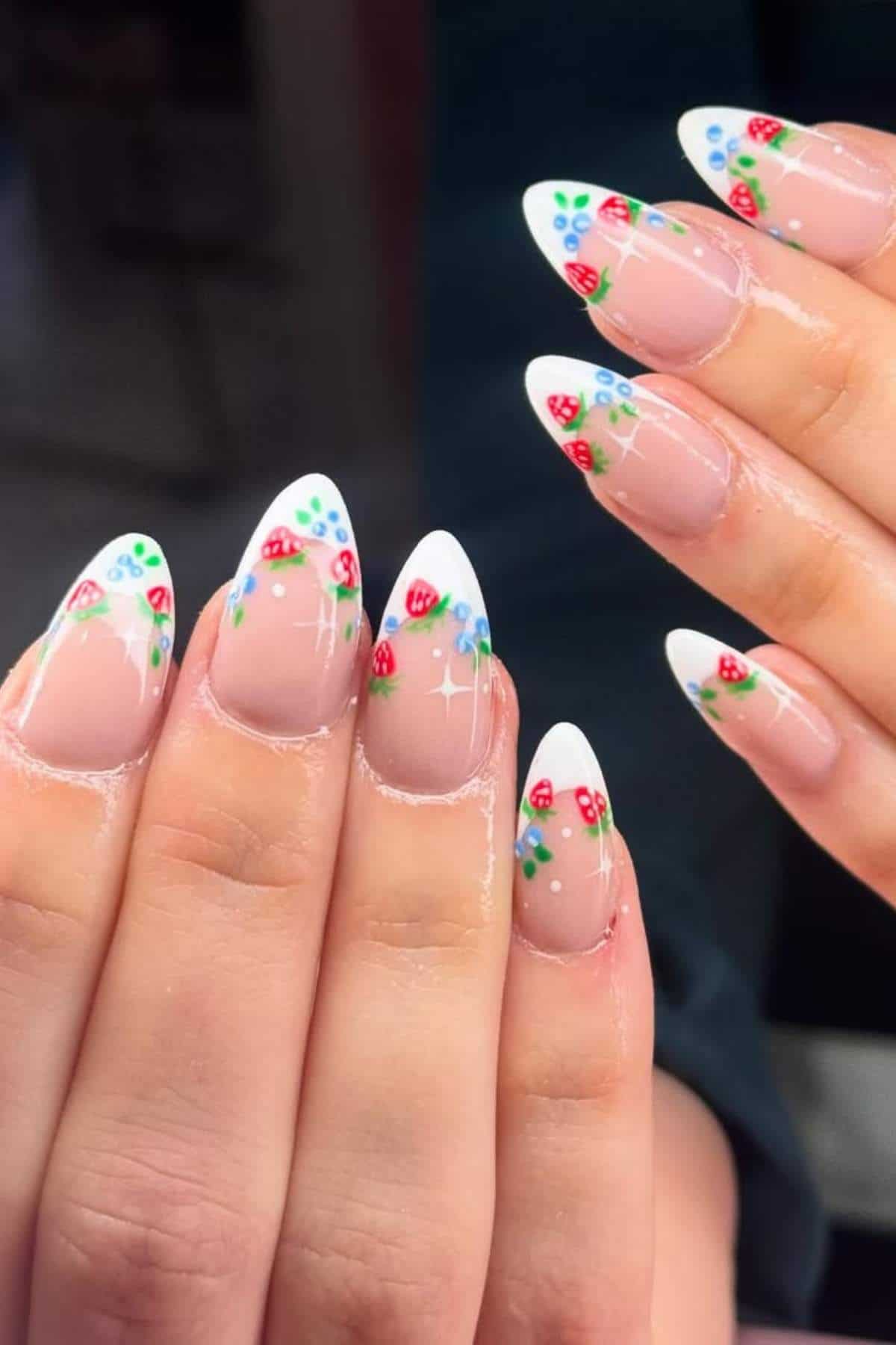 Close-up of hands with almond-shaped nails featuring a French tip design decorated with small red strawberries and green leaves.