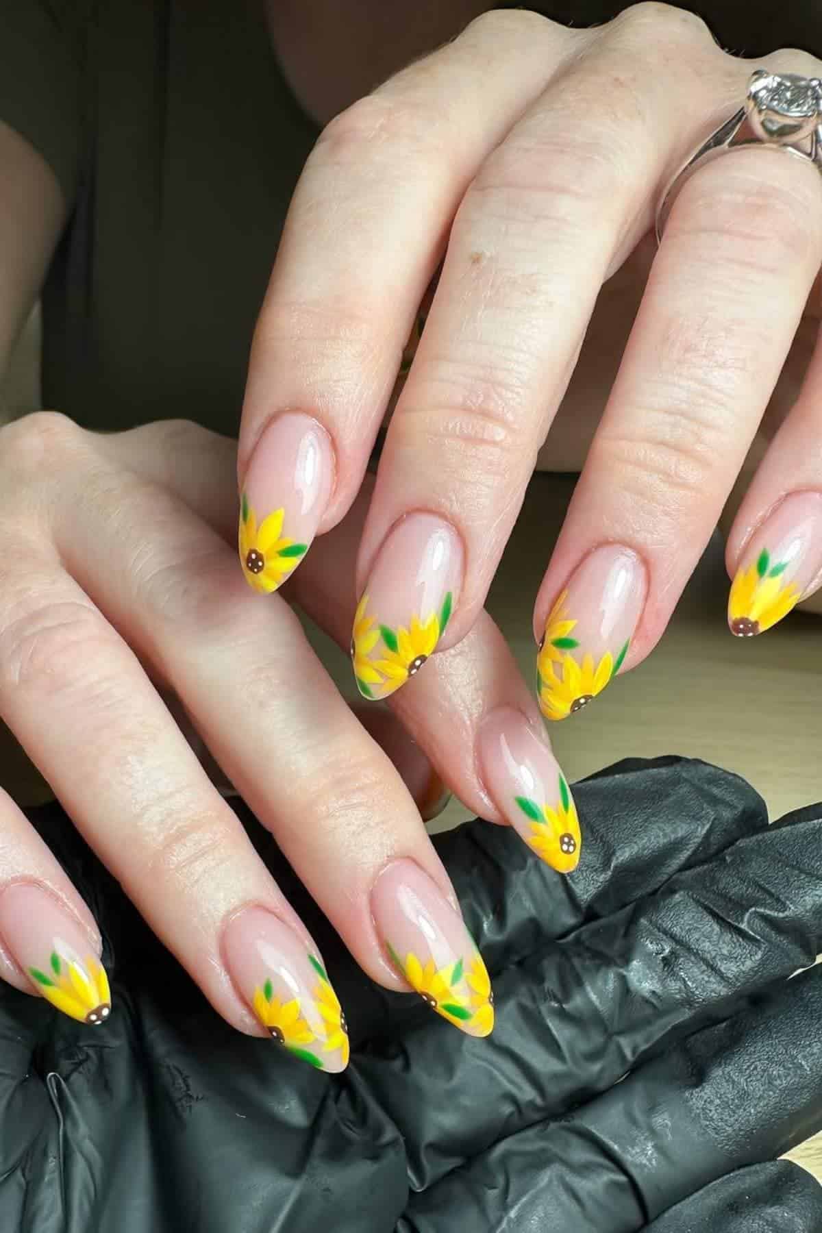 Hands with almond-shaped nails featuring yellow sunflower nail art, green leaves, and brown centers on a nude base. One hand wears a silver ring; a gloved hand is partially visible.