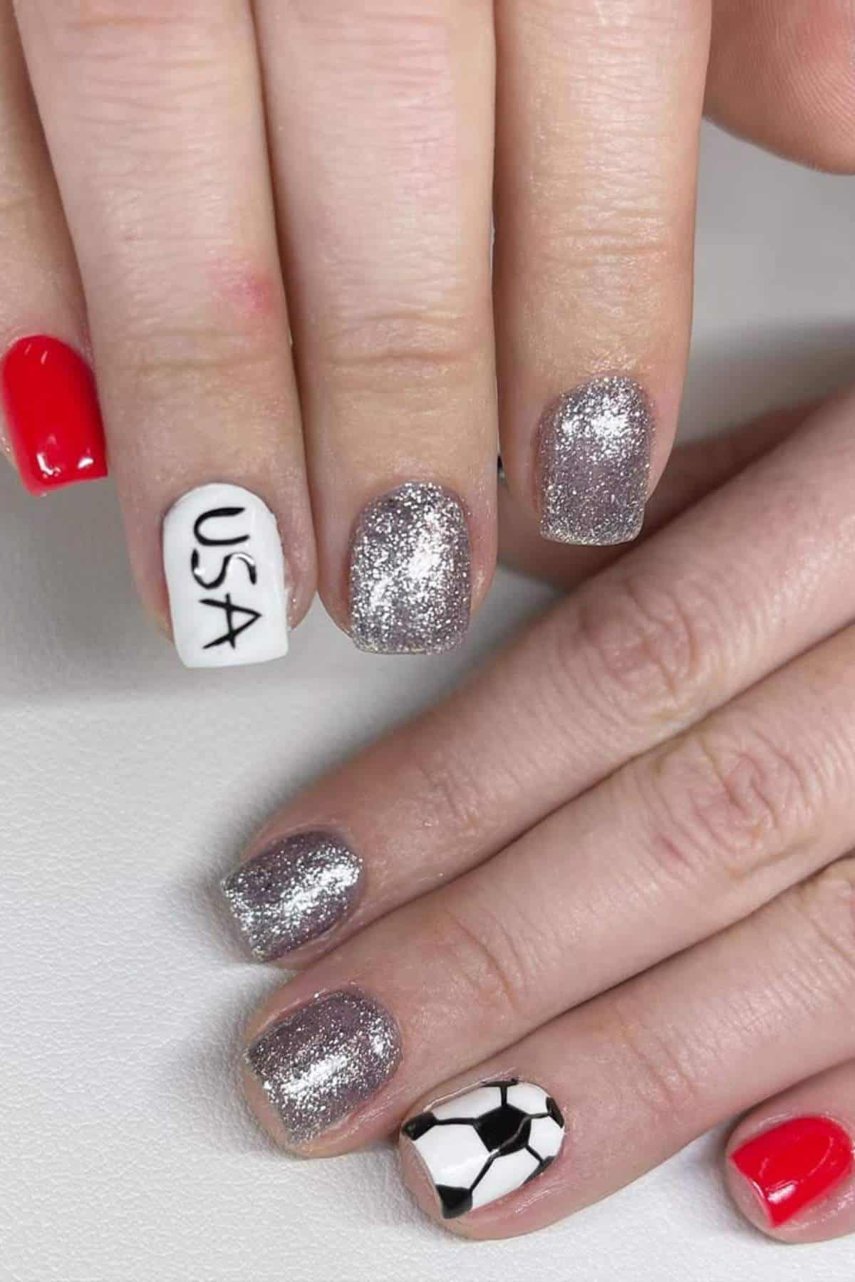 A hand with manicured nails featuring red, silver glitter, one white nail with "USA" text, and one nail painted like a soccer ball.