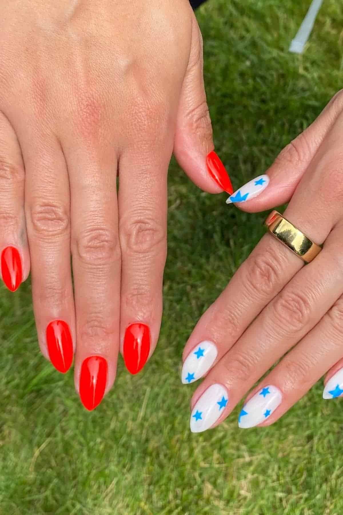 Two hands with manicured nails; one has solid red polish, the other has white polish with blue star designs. The person is wearing a gold ring on one finger. The background is grass.