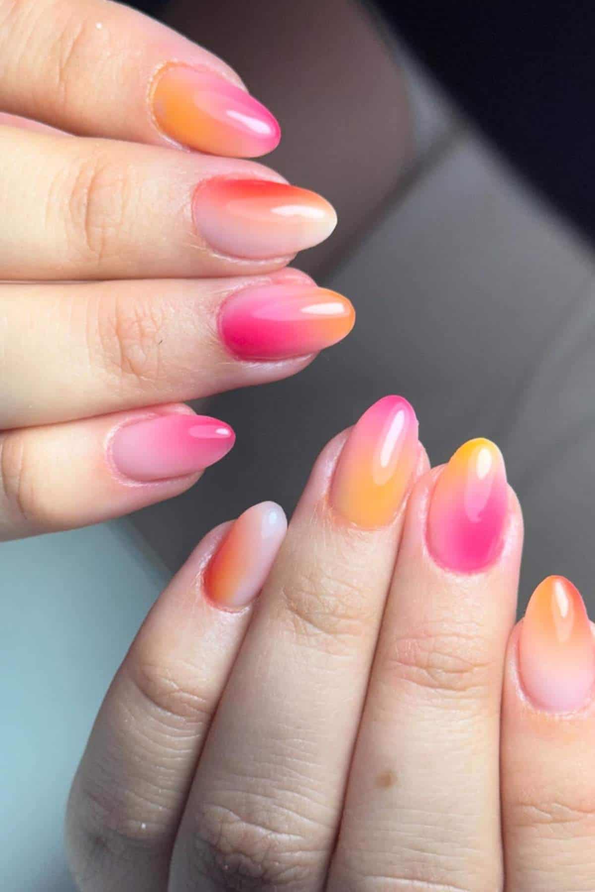 Close-up of hands with oval-shaped nails painted in a gradient ombre of pink, orange, and yellow shades.
