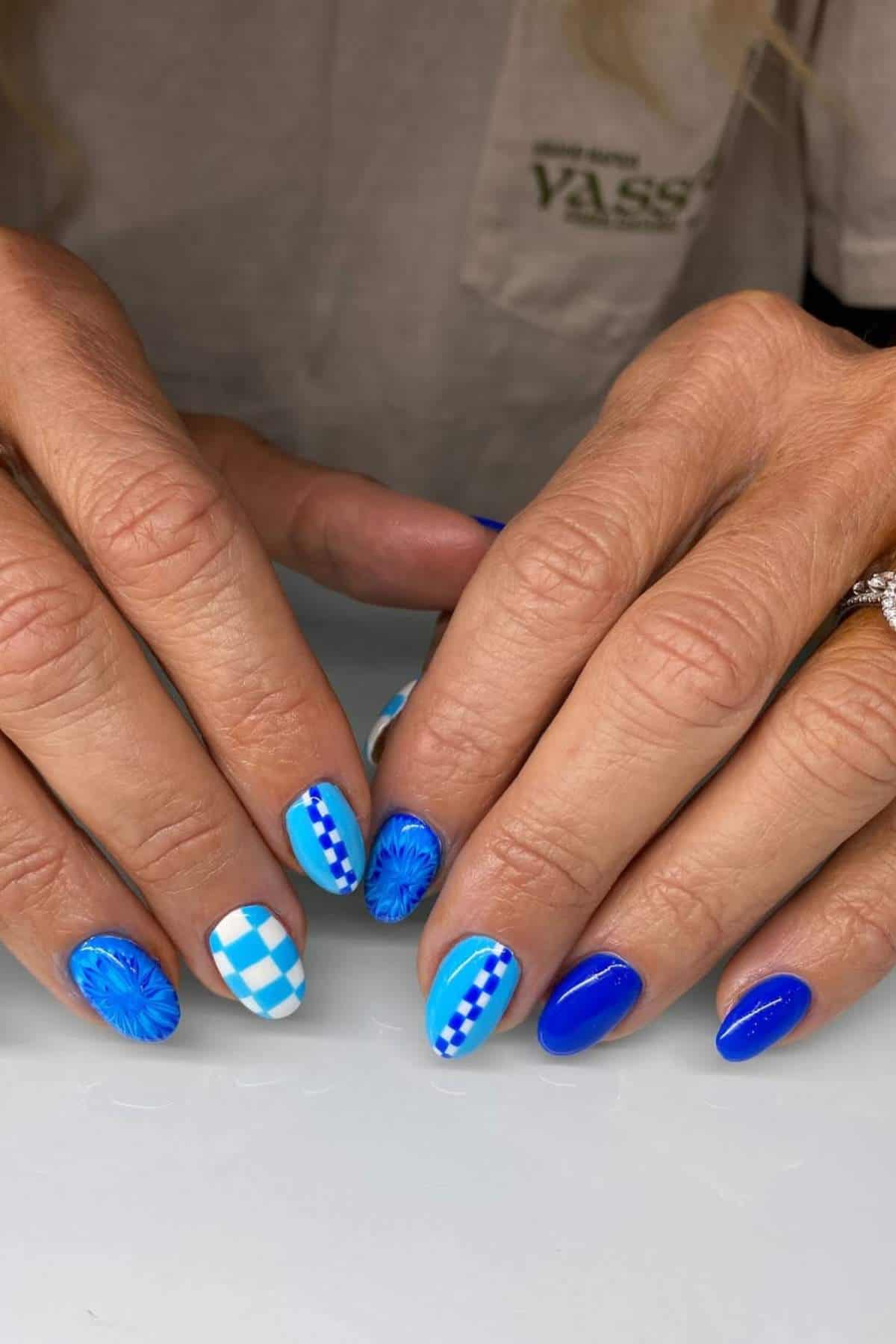 Close-up of hands with blue-themed nail art featuring checkered, striped, and abstract patterns on short nails.