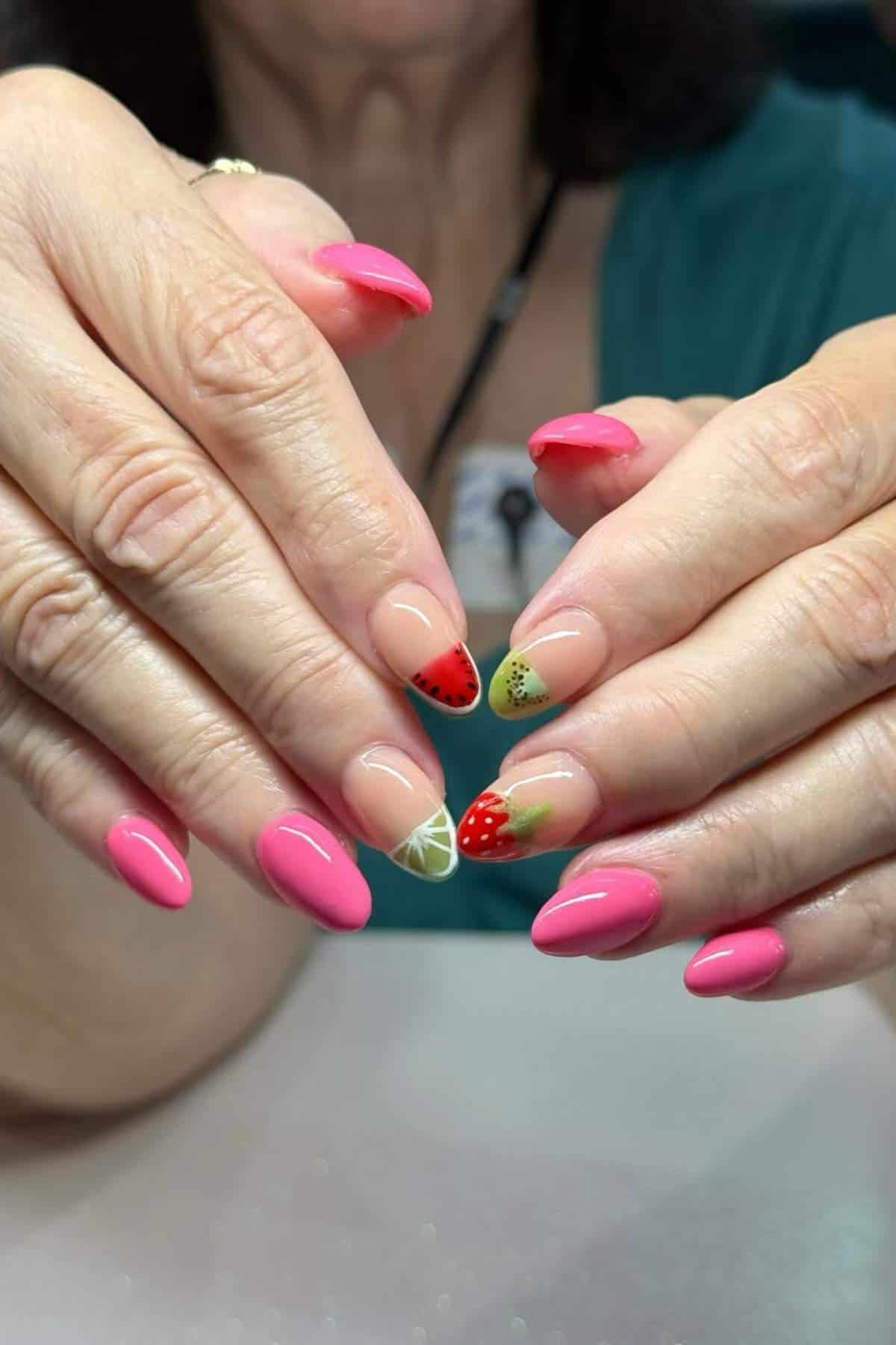 Hands with long nails painted pink and decorated with fruit designs, including strawberries and limes, on a few accent nails.
