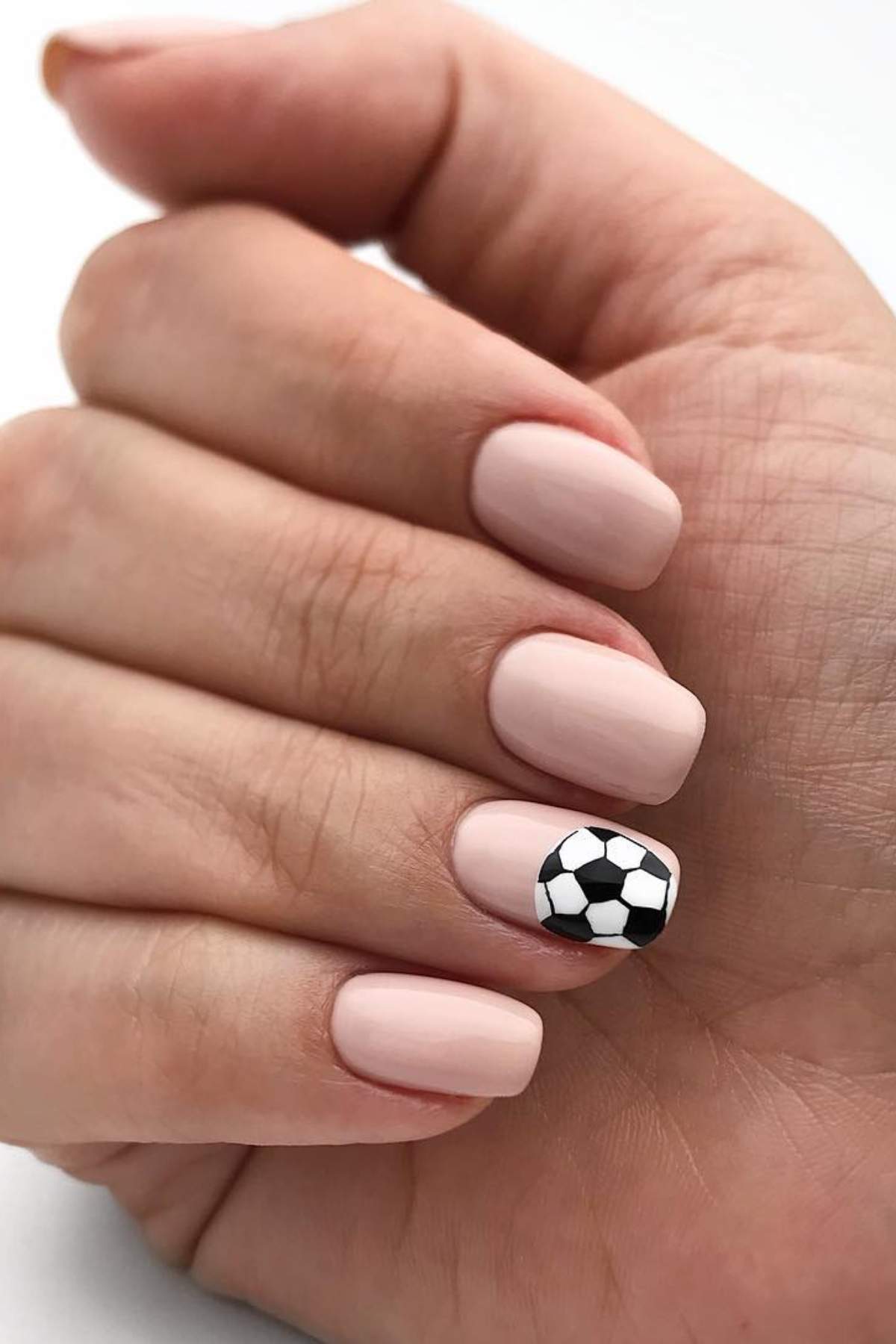 A hand with pale nude nail polish on all nails except the pinky, which features a black and white soccer ball design.