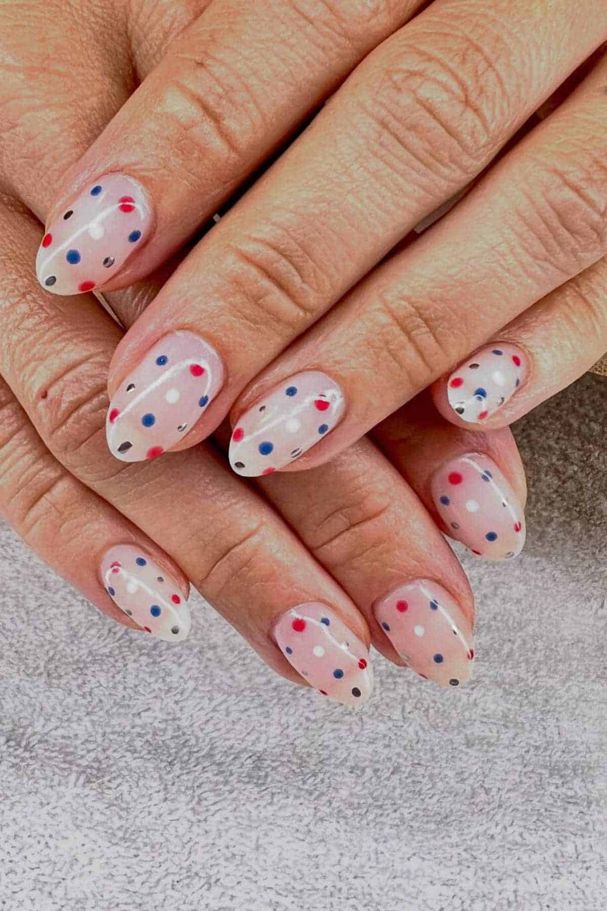 Hands with almond-shaped nails featuring a clear base and multicolored polka dot nail art in blue, red, and black.