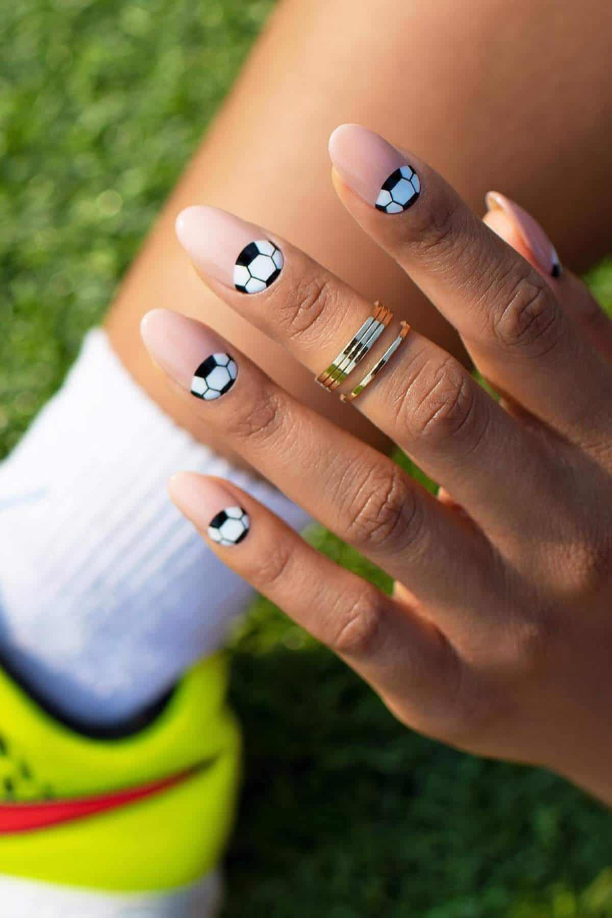 A hand with soccer ball nail art rests on a knee; person wears a white sock, yellow sneaker, and gold rings. Grass is visible in the background.