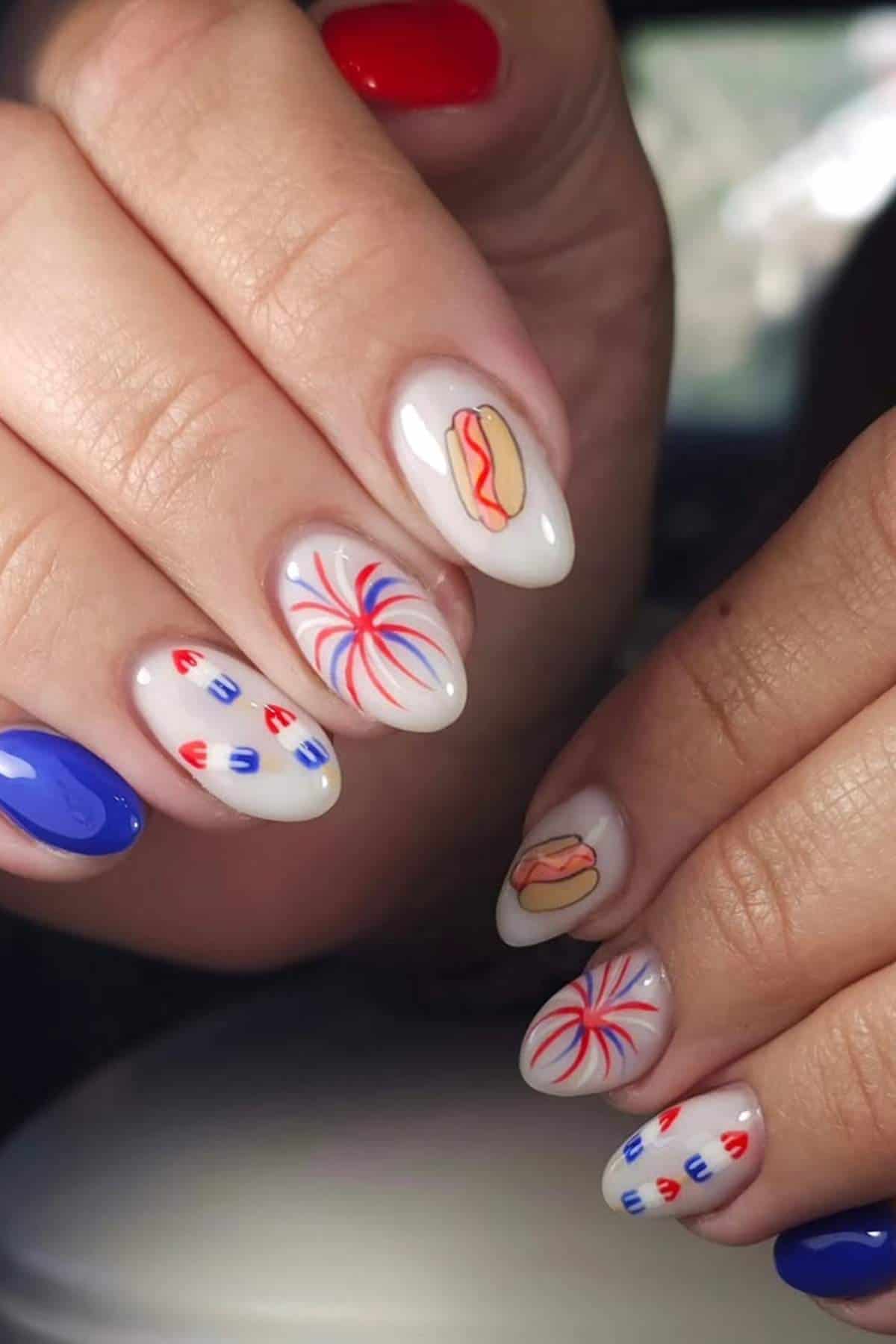 Hands with almond-shaped nails featuring patriotic red, white, and blue designs, fireworks, hot dogs, and solid red and blue accent nails.