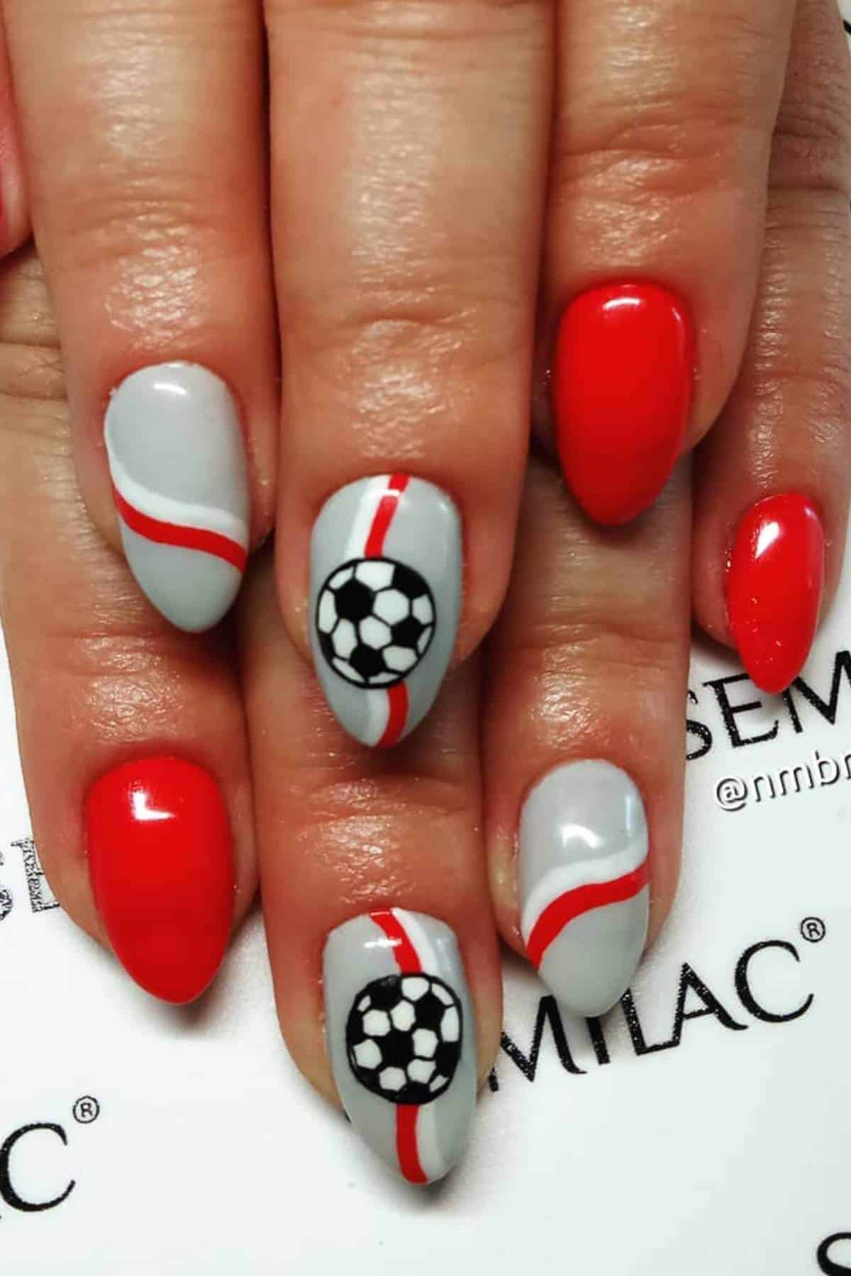 A hand with manicured nails painted red and gray; two nails feature a soccer ball design and red and white diagonal stripes.