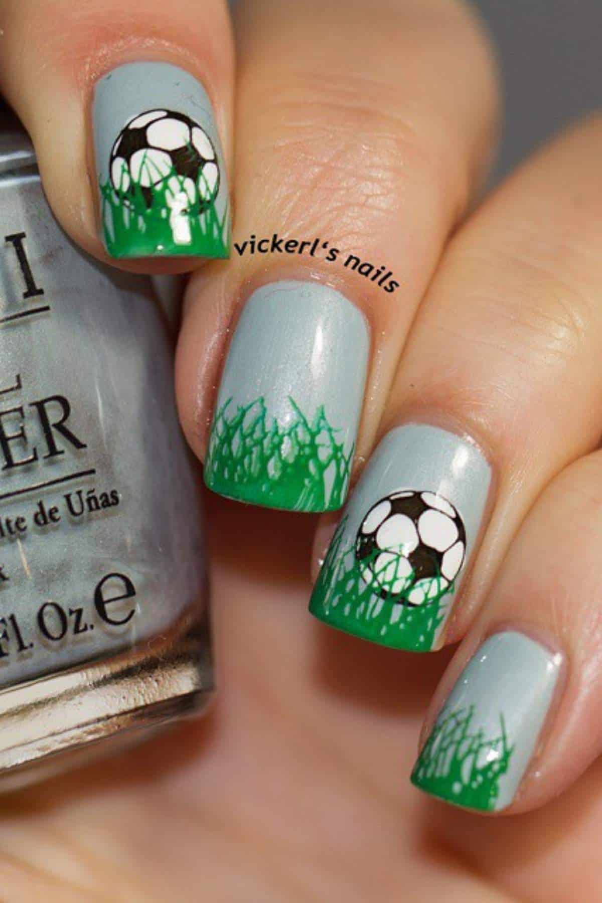 Fingernails painted with a green grass design and soccer balls on a light gray base, holding a nail polish bottle.