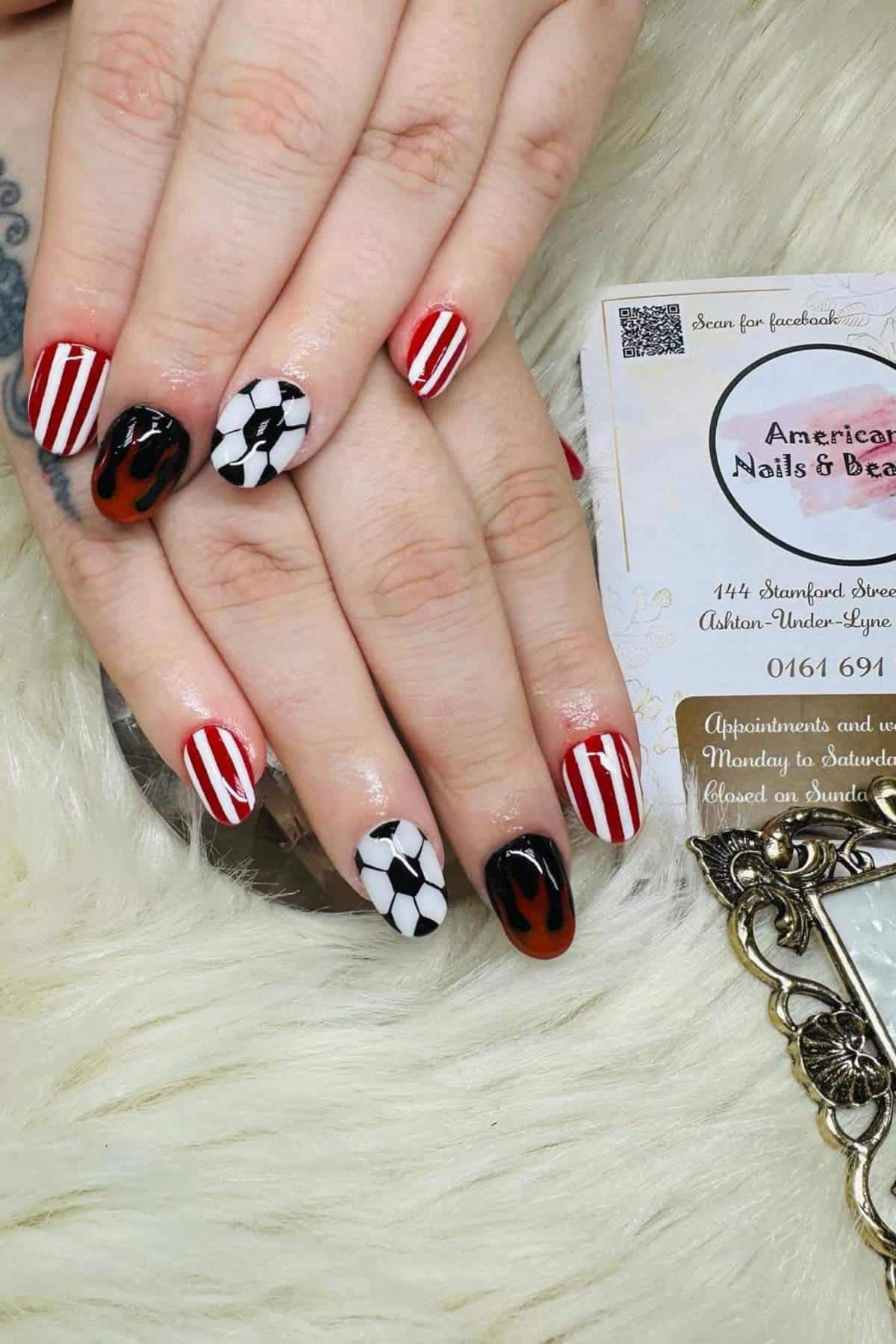 A hand with manicured nails featuring red and white stripes, black and white soccer balls, and black flames on a light fur surface, next to a business card and a small mirror.