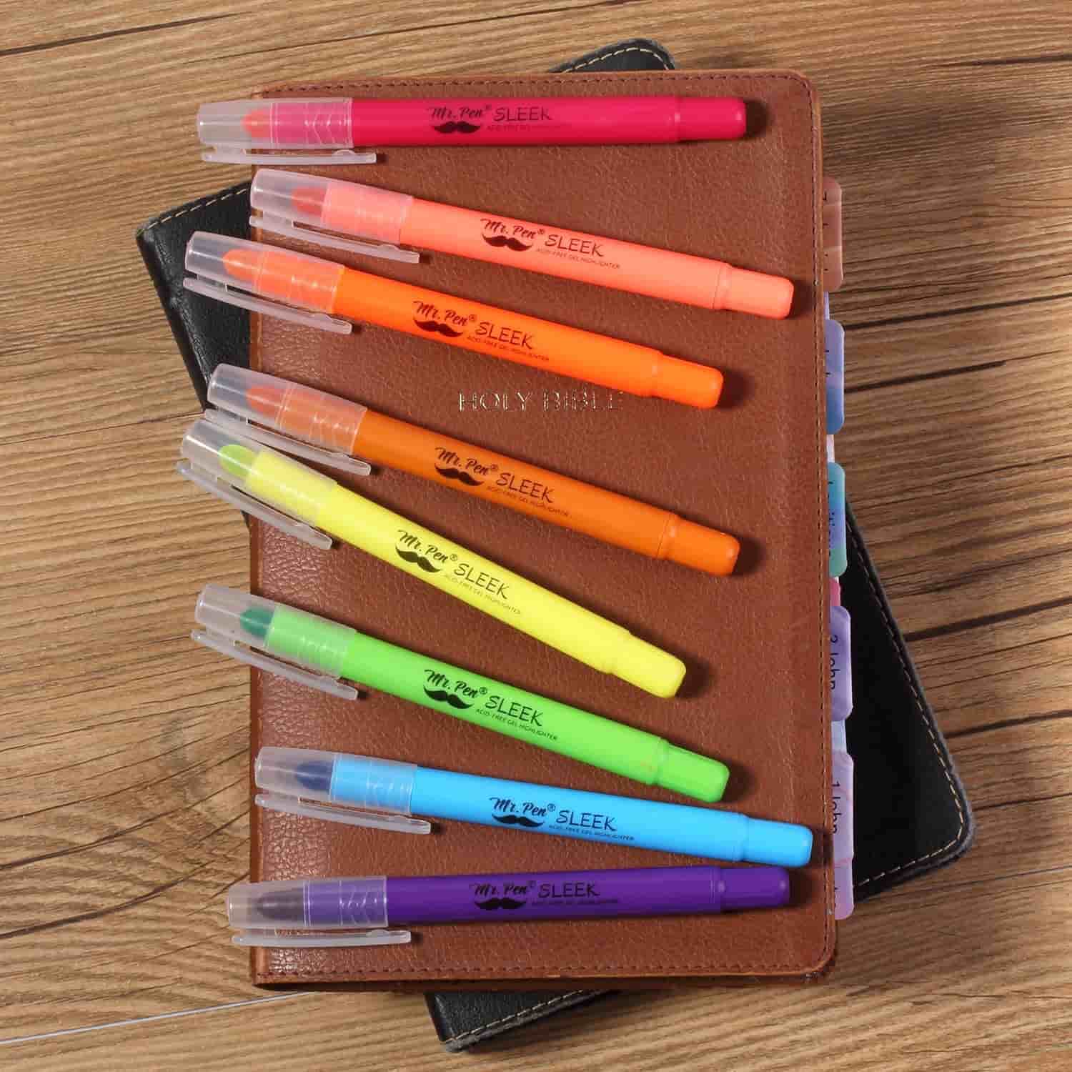 Eight colorful highlighters are placed diagonally on a closed brown leather-bound book with tabbed dividers, all resting on a wooden surface.