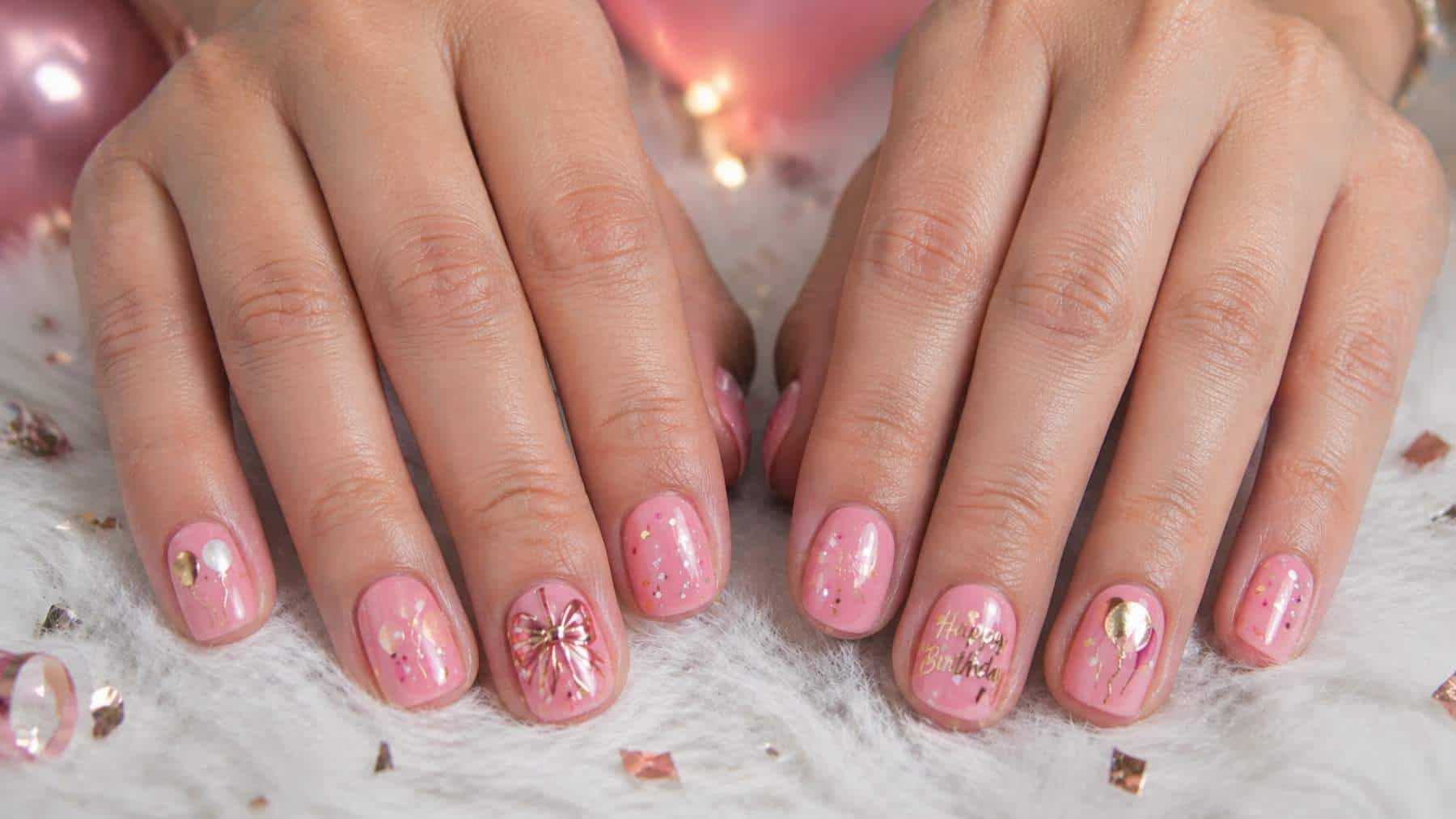 Close-up of hands with short, round nails painted pink and decorated with gold designs, including bows, balloons, and glitter accents.