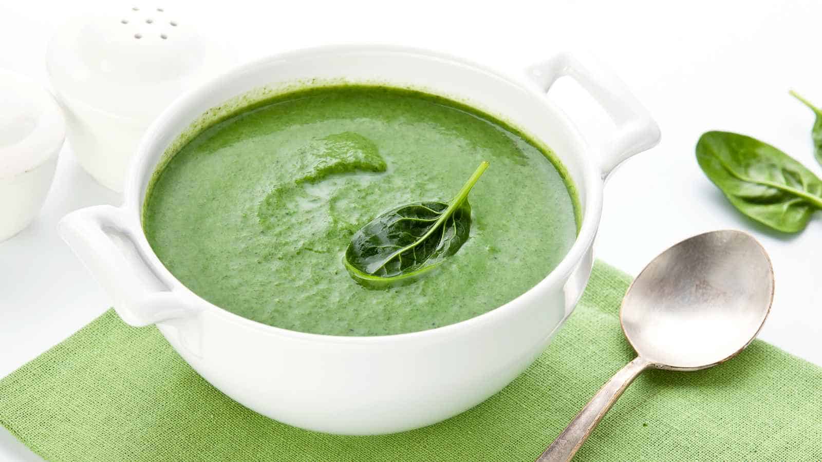 A white bowl of green spinach soup, garnished with a spinach leaf, sits on a green napkin next to a silver spoon.