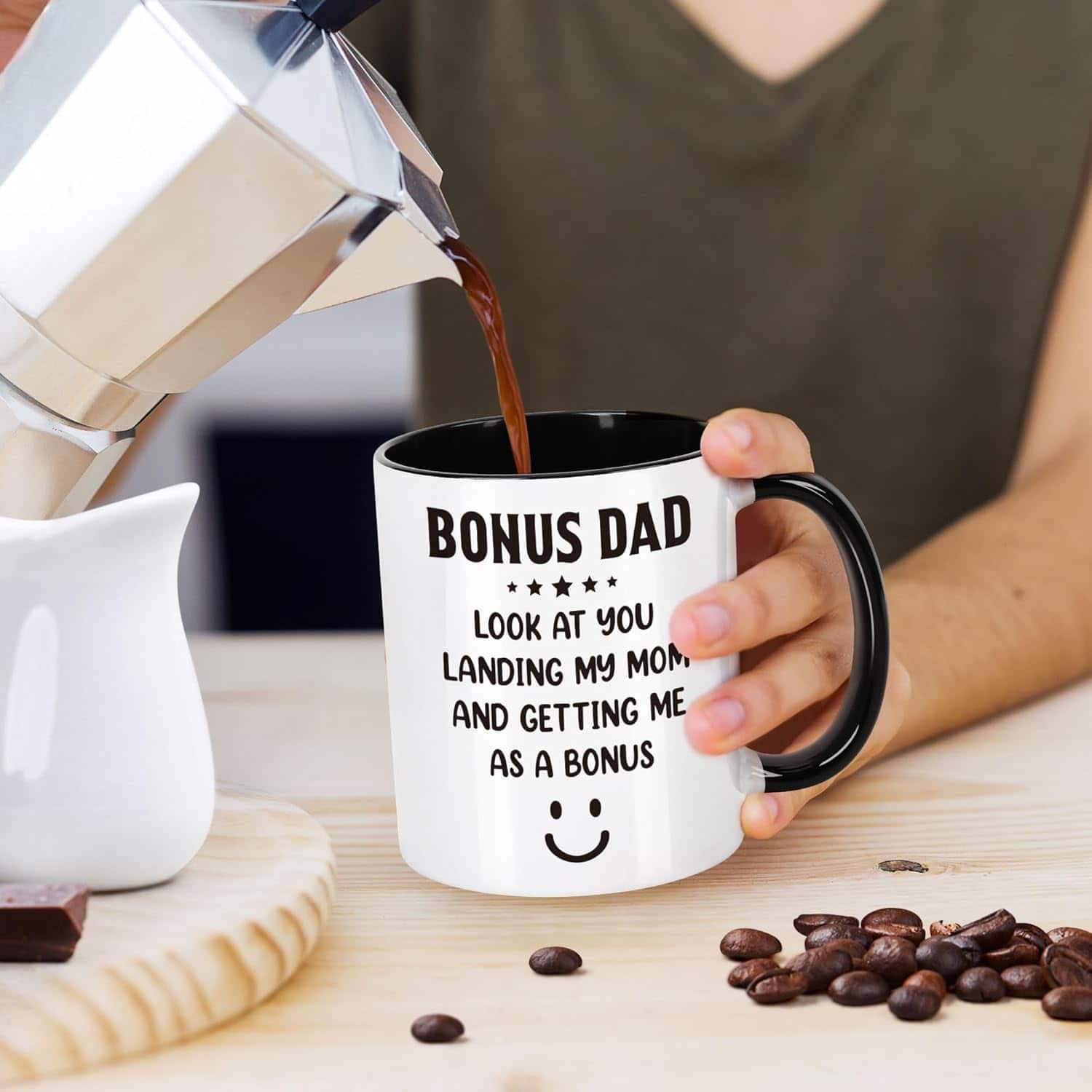 A person pours coffee into a mug that reads, "Bonus Dad. Look at you landing my mom and getting me as a bonus," with a smiley face. Coffee beans and a chocolate piece are on the table.