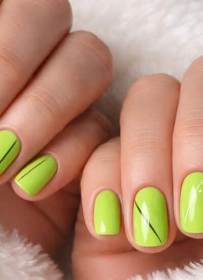 Hands with short, neatly manicured nails painted bright lime green, featuring thin black lines and small white dot accents, resting on a white textured fabric.
