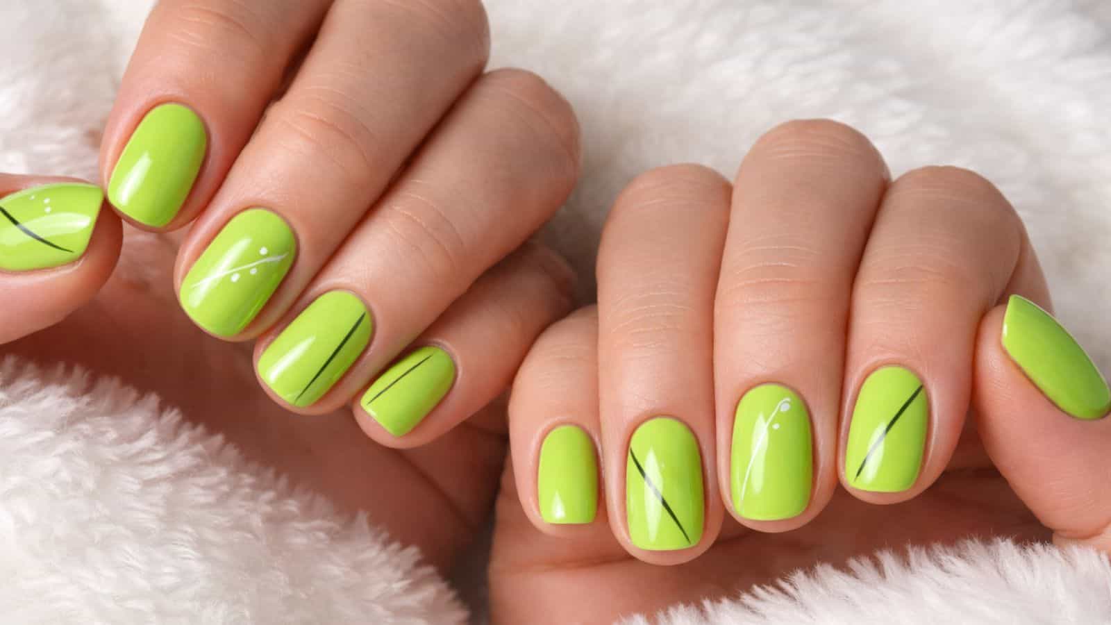 Hands with short, neatly manicured nails painted bright lime green, featuring thin black lines and small white dot accents, resting on a white textured fabric.