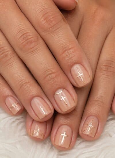 Close-up of hands with short, nude-colored nails featuring minimalist gold and white cross designs on a soft white textured surface.