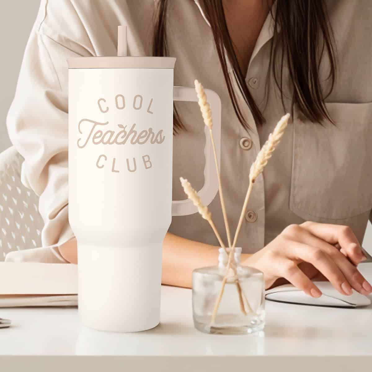 A woman sits at a desk with a notebook, pen, and a large white tumbler labeled "Cool Teachers Club" beside a small vase holding dried wheat stalks.