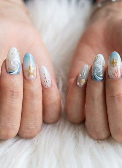 Close-up of hands with long, almond-shaped nails featuring ocean-themed nail art, including waves, shells, starfish, anchors, and pastel blue and white accents.