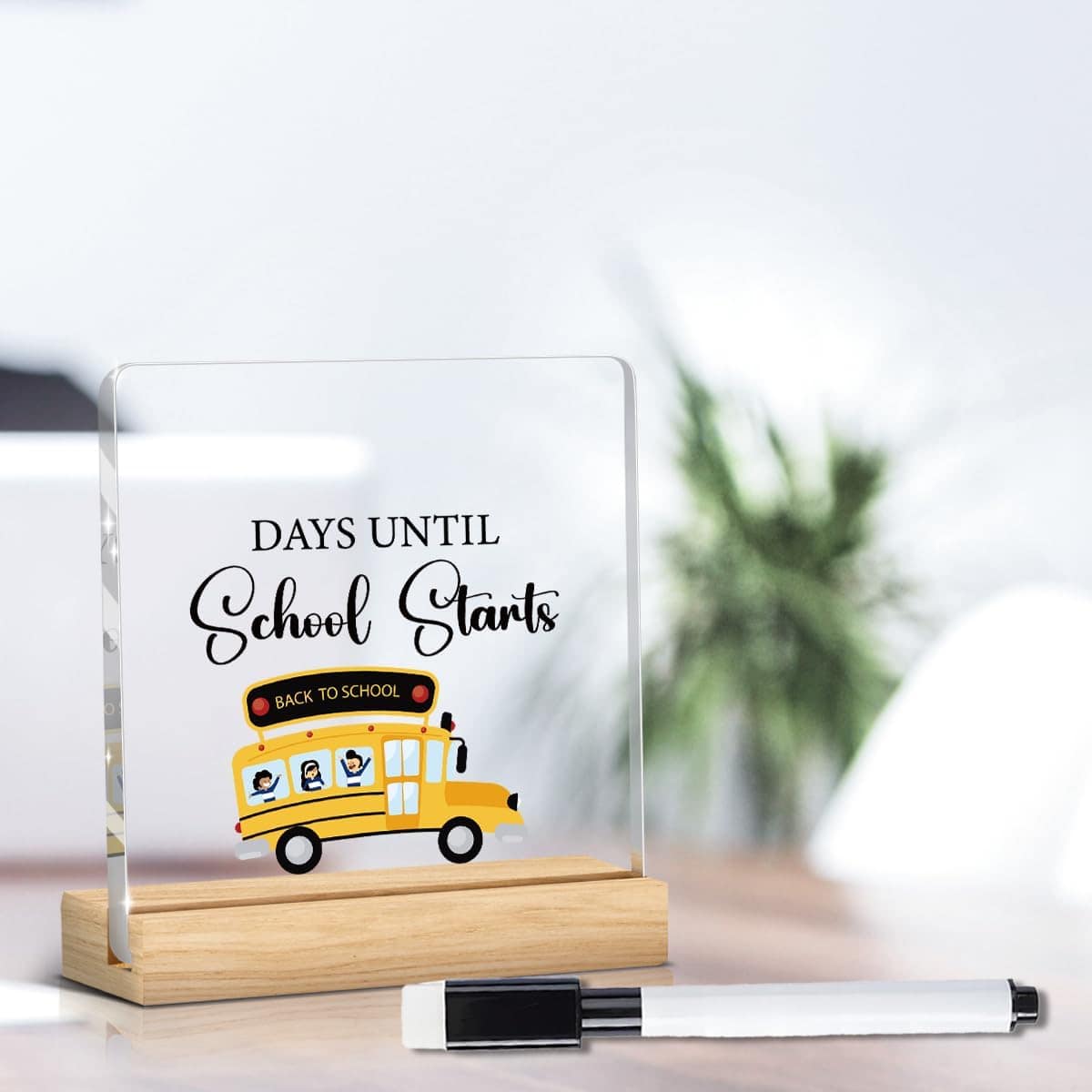 A clear acrylic sign with "Days Until School Starts" and a yellow school bus illustration, placed on a wooden stand next to a dry erase marker and a plant in the background.