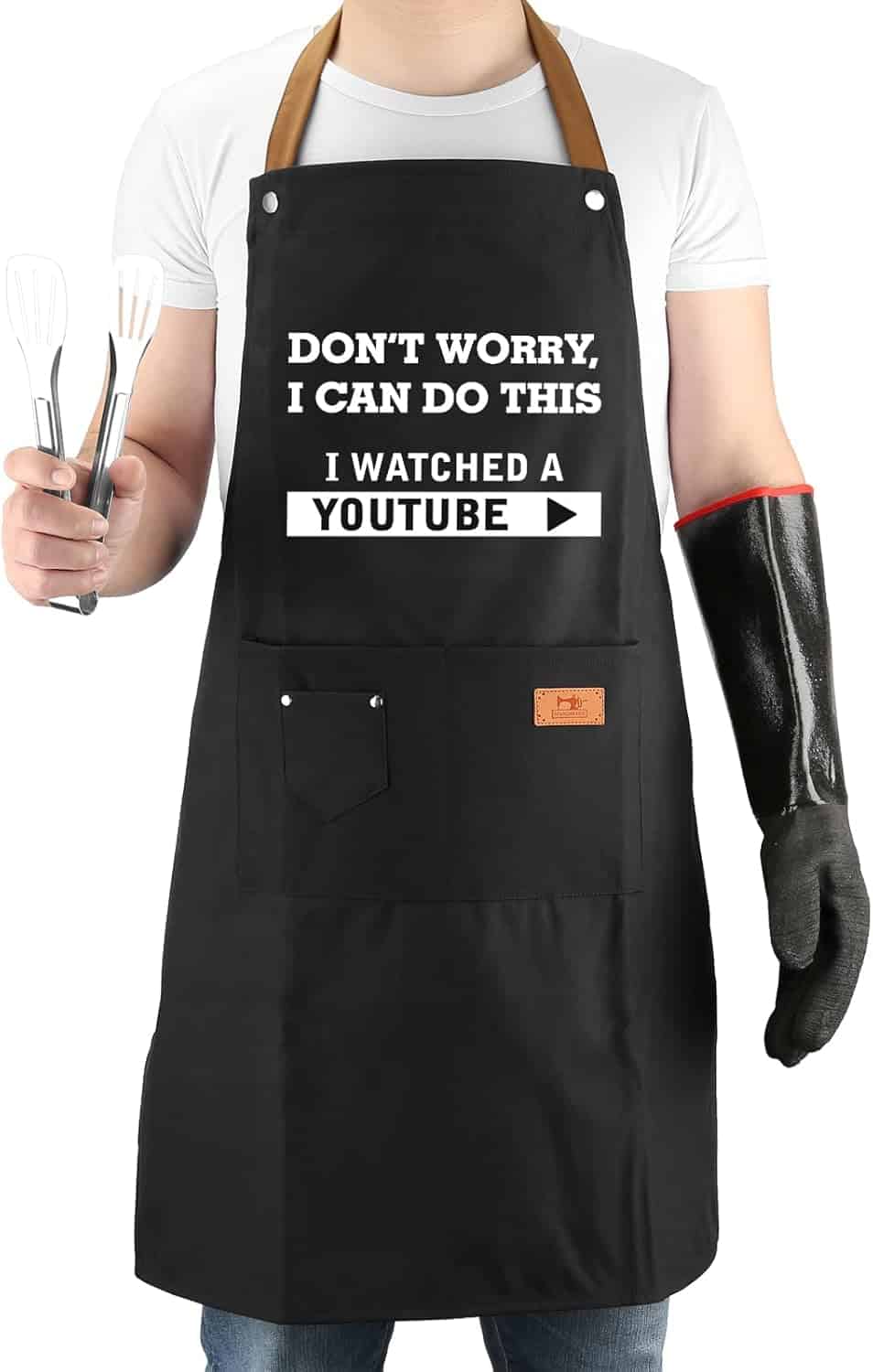 Person wearing a black apron that says, "DON'T WORRY, I CAN DO THIS. I WATCHED A YOUTUBE," holding two spatulas in one hand and wearing a black glove on the other hand.