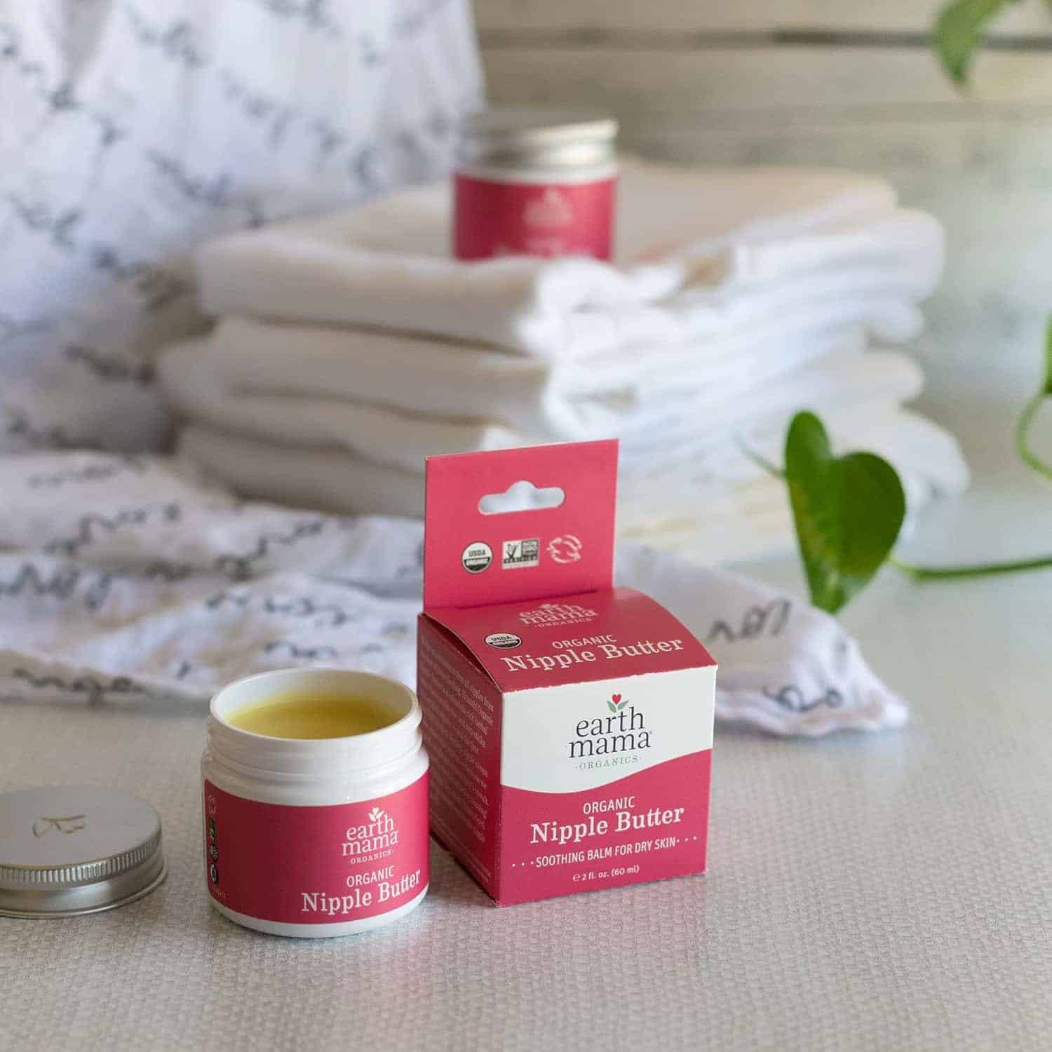 Open jar of Earth Mama Organic Nipple Butter with its box in front of folded white cloths on a table, plant in the background.