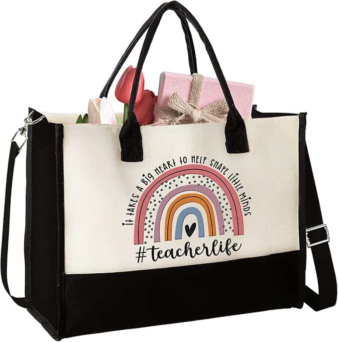 Canvas tote bag with black handles, rainbow design, and text "#teacherlife"; bag contains a pink gift box and some flowers.