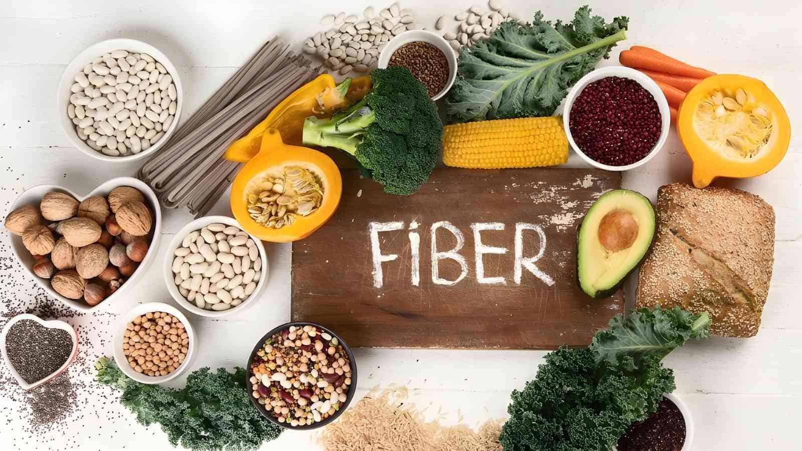 An assortment of high-fiber foods including beans, broccoli, whole grain bread, avocado, nuts, seeds, carrots, corn, and leafy greens surrounding a chalkboard labeled "FIBER.