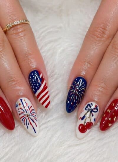 Hands with long almond-shaped nails painted in red, white, and blue with patriotic designs including stars, stripes, fireworks, cherries, and the American flag.
