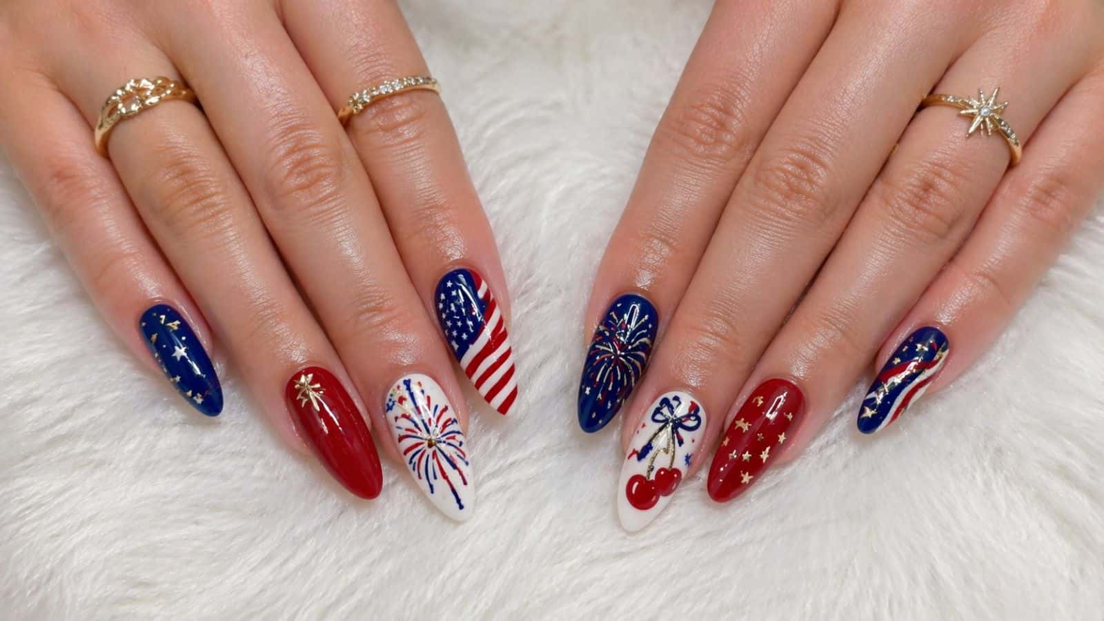 Hands with long almond-shaped nails painted in red, white, and blue with patriotic designs including stars, stripes, fireworks, cherries, and the American flag.