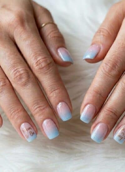 Close-up of two hands with short, square-shaped nails painted in a blue and white gradient with delicate designs and text on some nails. The person is wearing thin gold rings.