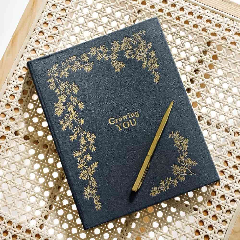 A dark blue journal with gold floral designs and the words "Growing YOU" on the cover, placed on a woven rattan surface next to a gold pen.