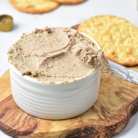 A knife spreading a creamy brown pâté from a white ramekin onto a cracker, with more crackers and a measuring spoon in the background.
