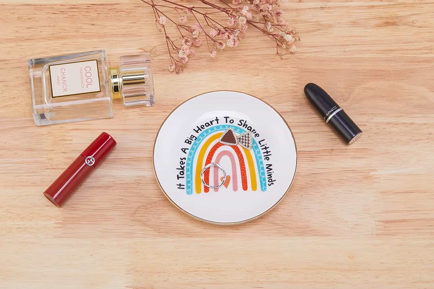 A decorative dish with the text "It takes a big heart to shape little minds" sits on a wooden surface, surrounded by perfume, red lipstick, black lipstick, and dried flowers.