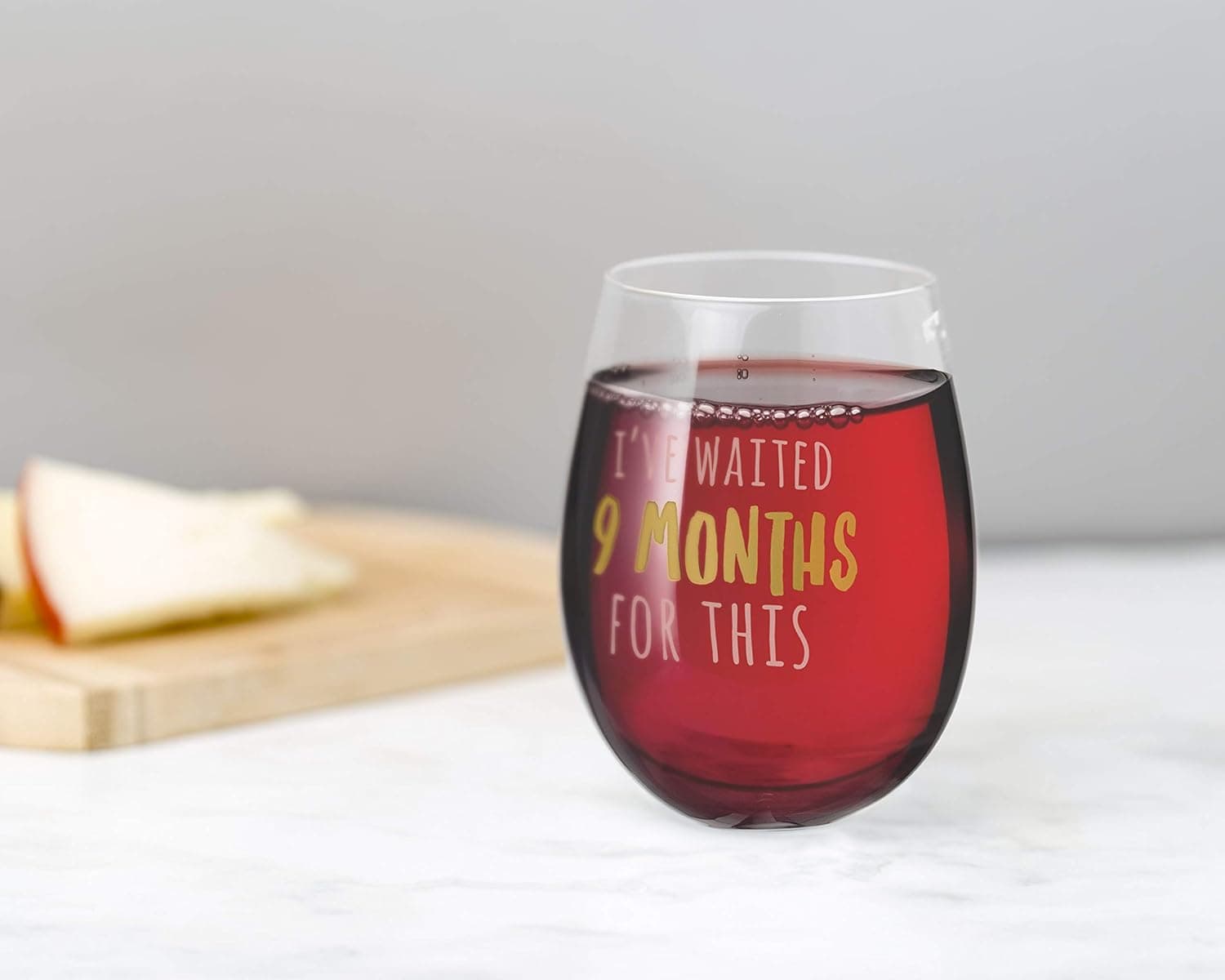 A stemless wine glass filled with red wine, featuring the phrase "I've waited 9 months for this;" cheese and apples are on a wooden board in the background.