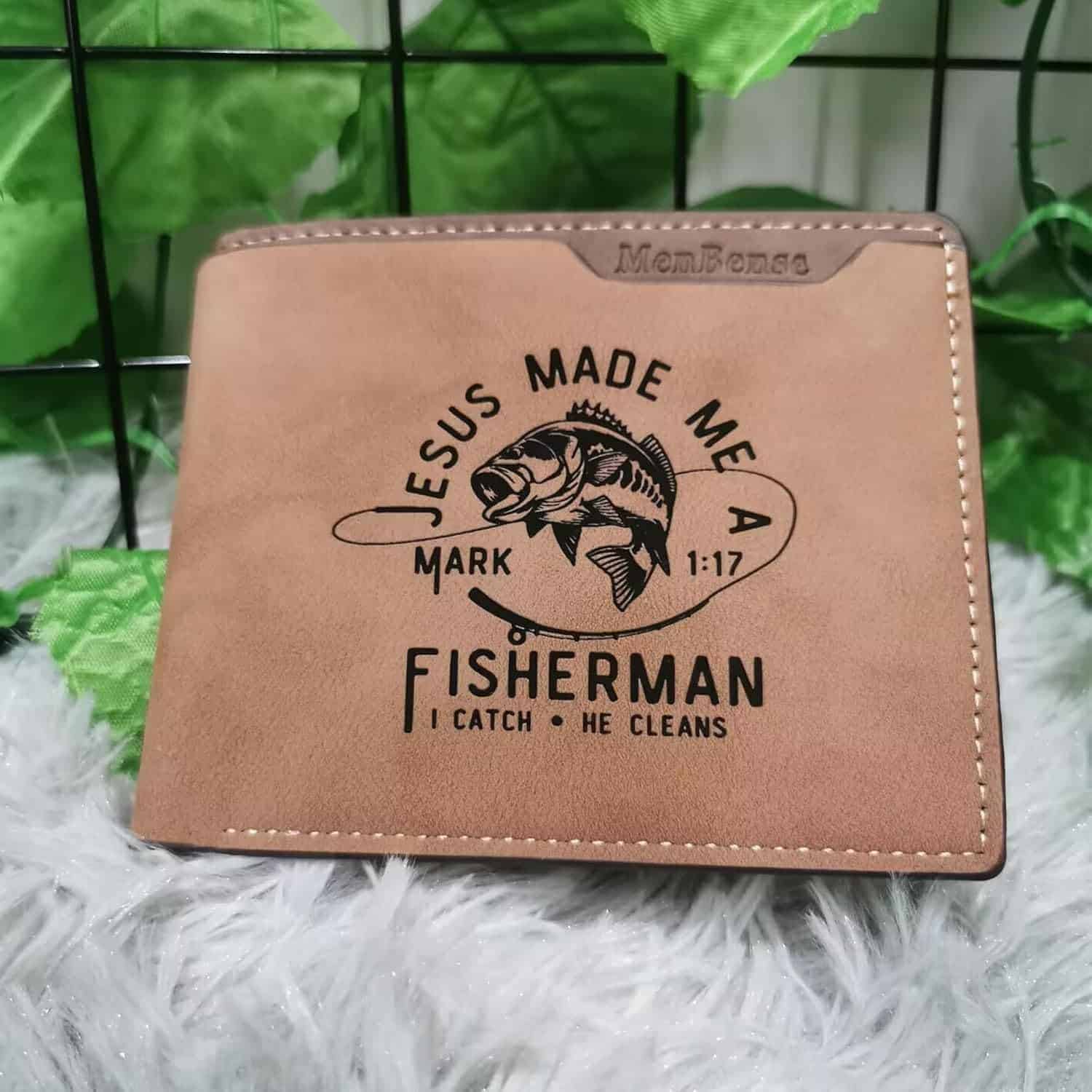 A brown wallet with the text "Jesus made me a fisherman, Mark 1:17, I catch, He cleans" and an image of a fish printed on it, placed on a furry surface with green leaves in the background.