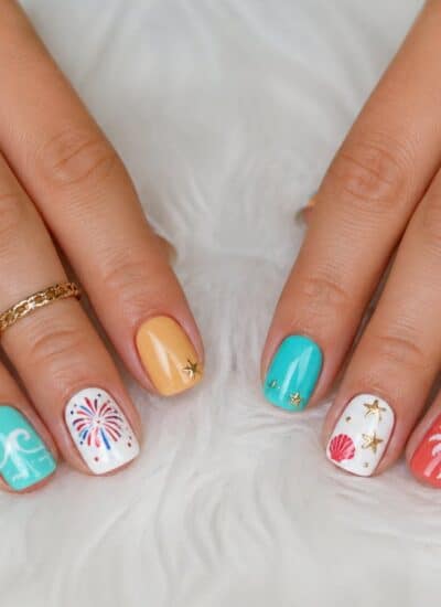 Hands with summer-themed nail art including palm trees, fireworks, starfish, and stripes in orange, blue, and white, shown on a white textured surface.