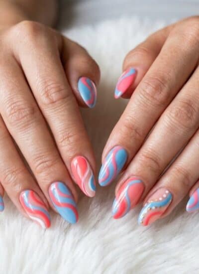 Two hands with long nails featuring a pink and blue wavy design and an accent nail with small white dots, resting on a white furry surface.