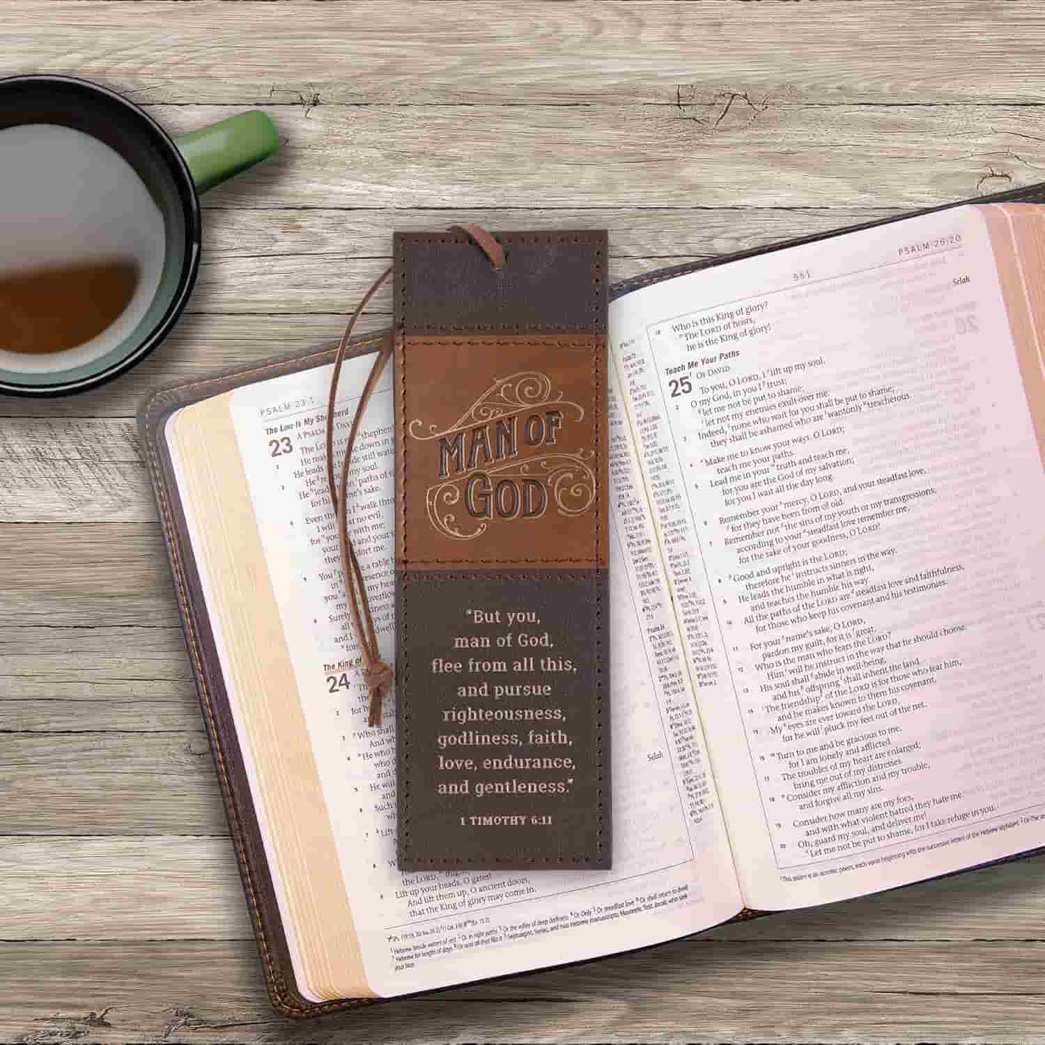 An open Bible lies on a wooden surface with a leather "Man of God" bookmark and a cup of coffee nearby.