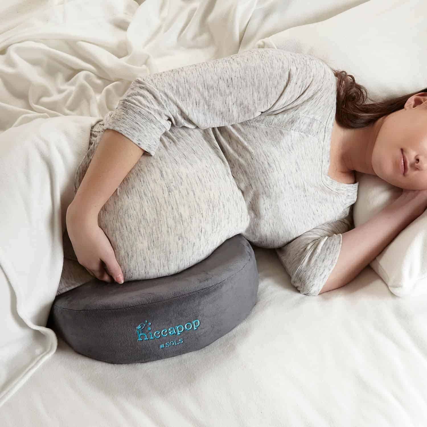 A pregnant woman lying on her side in bed uses a gray hiccapop pregnancy pillow for support under her belly.