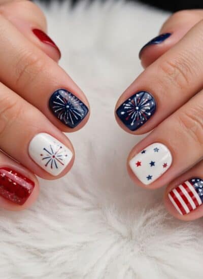 Hands with manicured nails featuring red, white, and blue designs, including stars, stripes, and fireworks, displayed on a white, furry surface.