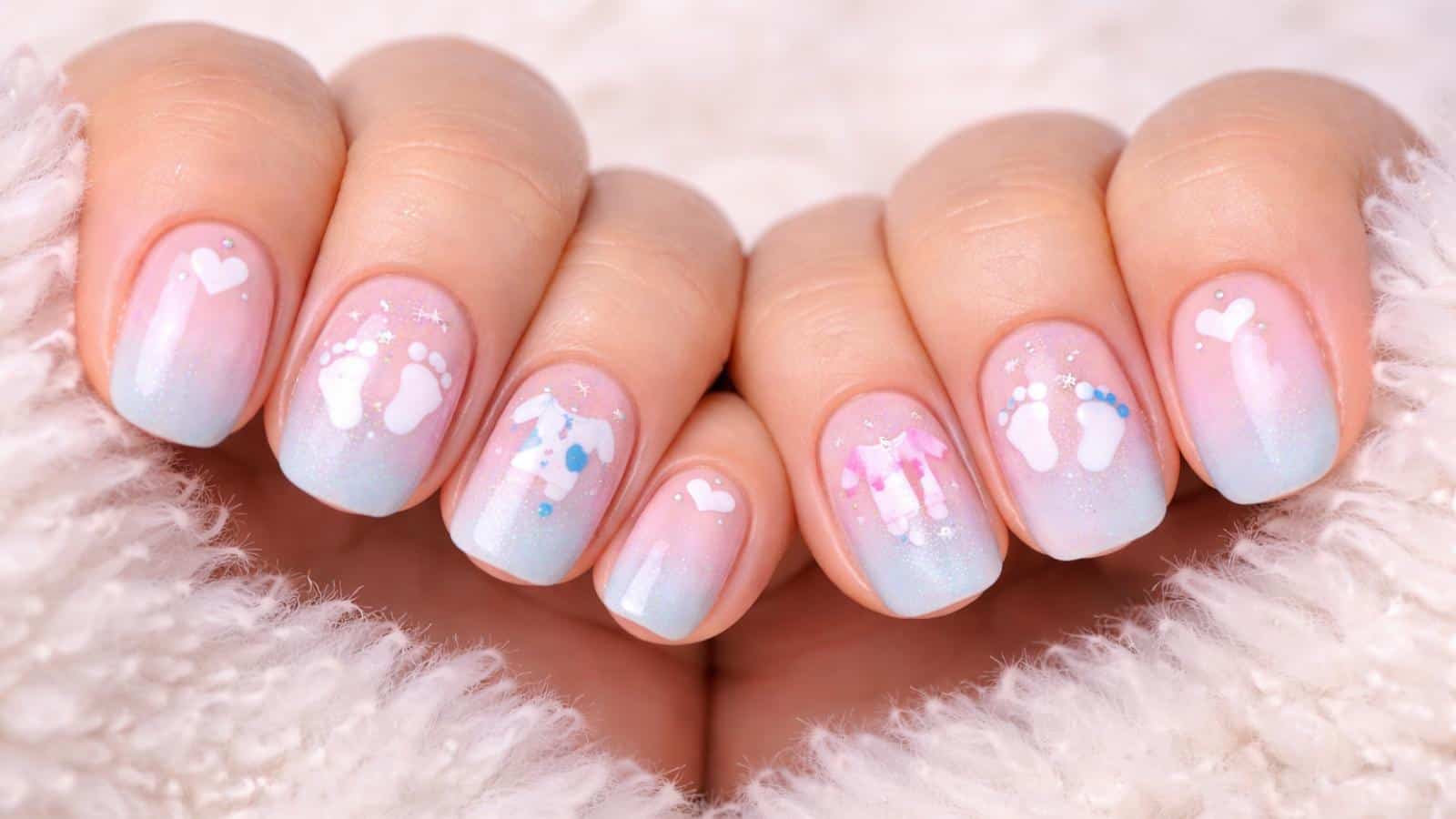Close-up of hands with pastel ombre manicured nails featuring white hearts, baby footprints, and baby clothes nail art designs.