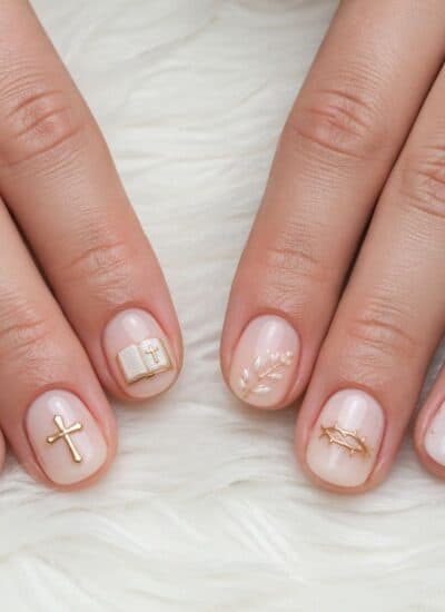 Close-up of hands with short, pale pink manicured nails decorated with gold religious-themed charms and symbols, displayed on a white fluffy surface.