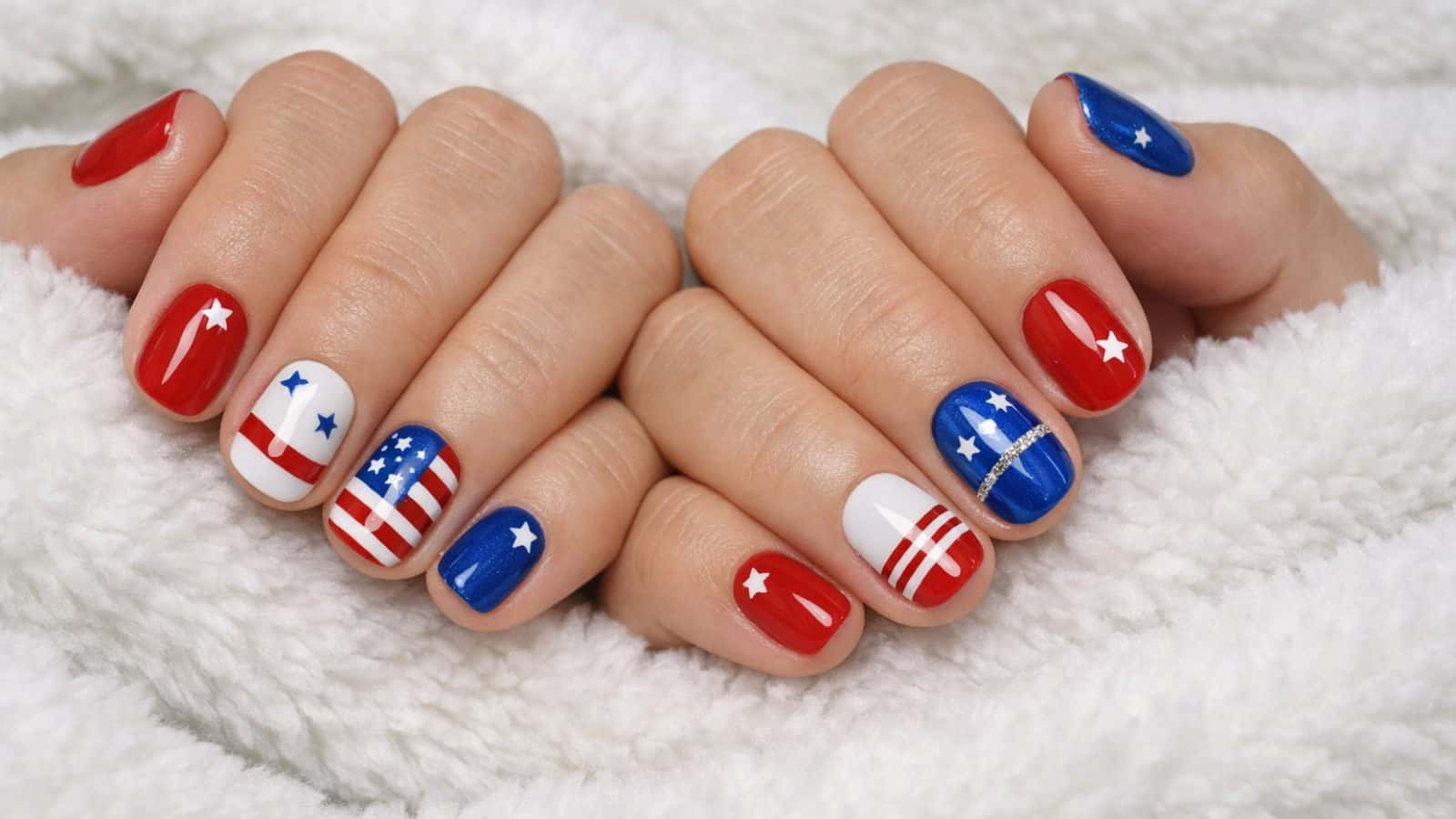 Hands with manicured nails painted in red, white, and blue with star and stripe patterns, resting on a white textured fabric.