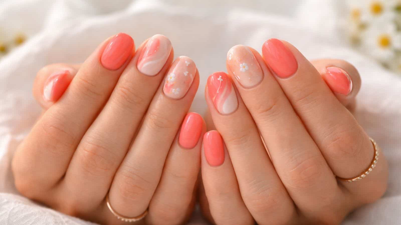 Hands with manicured nails painted in coral and white shades with floral and swirl designs, shown against a soft, light background.